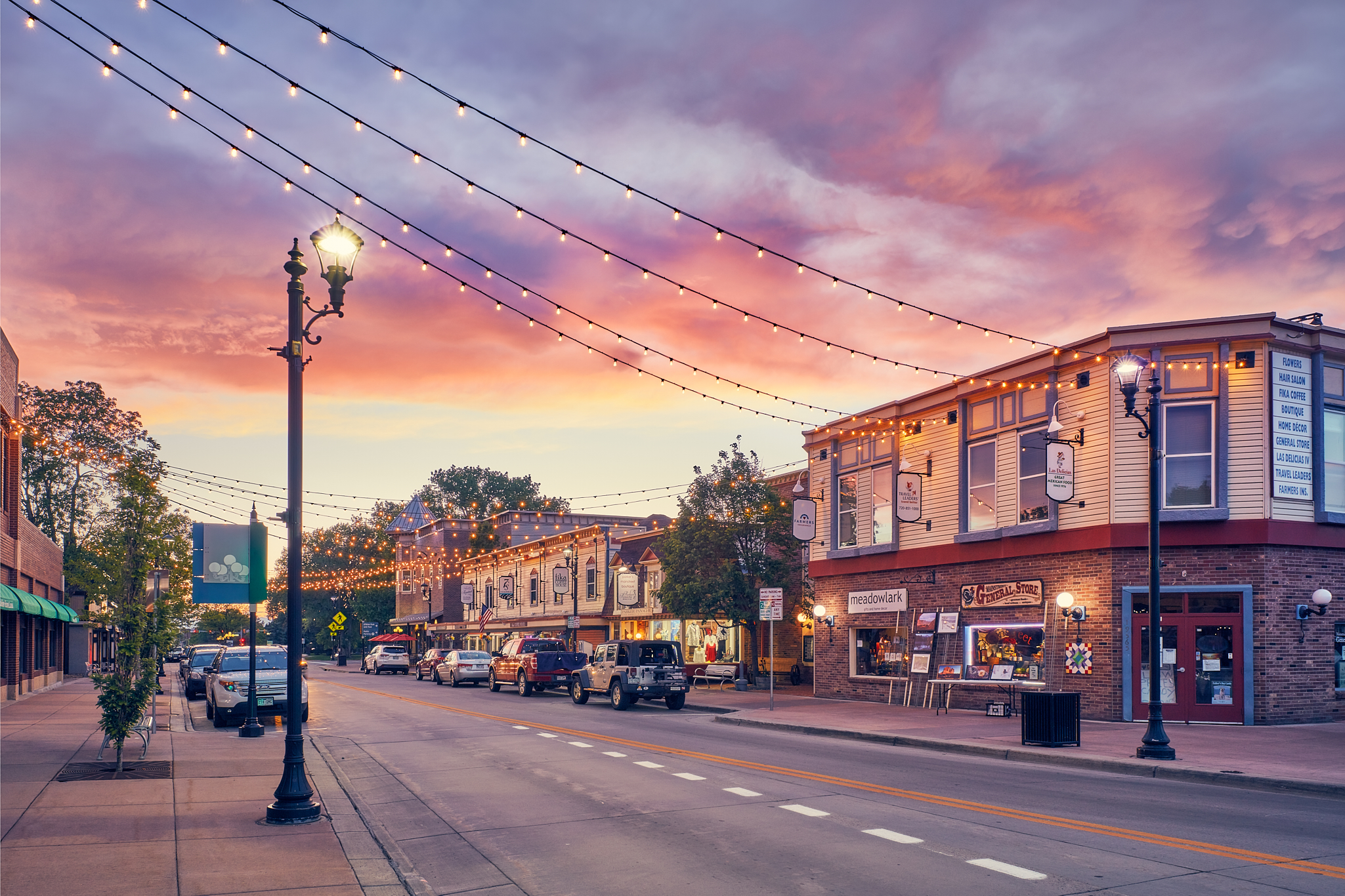 A city street in Parker, Colorado, featuring shops and cars, illuminated by a warm sunset glow. Great place for homeowning. Get a quote from Robert Castle Mortgage today.